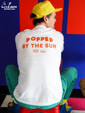 Person wearing a white shirt with 'Popped by the Sun' text, sitting on a colorful stool against a red and blue wall.